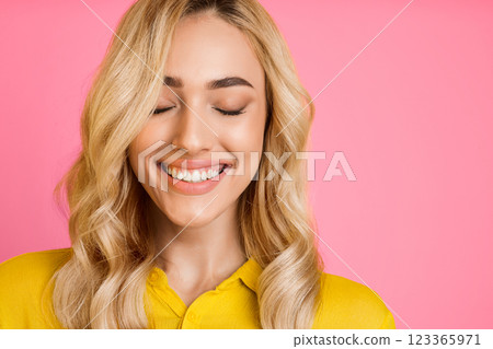 A young woman with long blonde hair smiles while looking down. She is wearing a yellow shirt and has her eyes closed. The background is a bright pink. A young woman with long blonde hair smiles while looking down. She is wearing a yellow shirt and has her eyes closed. The background is a bright pink. 123365971