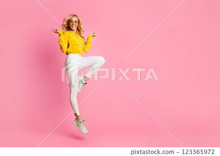 A young woman in a yellow hoodie and white jeans is jumping in the air with a big smile on her face. Her arms are outstretched and she is wearing sunglasses, copy space 123365972