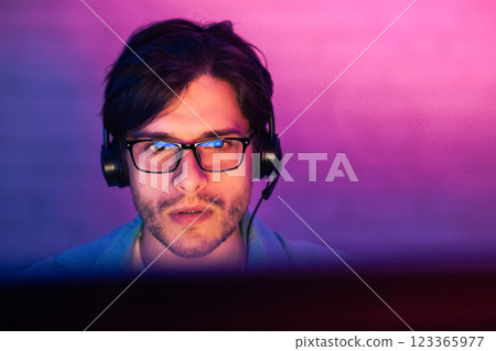 A young man with dark hair and a beard is wearing glasses and headphones, focused on a computer screen. The room is lit with a purple and pink gradient light, creating a vibrant and modern aesthetic 123365977