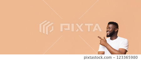 An African American man with a beard smiles and points to the left side of the frame. He is wearing a white t-shirt and standing against a tan background, copy space 123365980