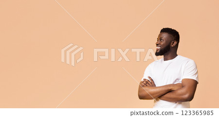 A smiling black man stands against a peach background with his arms crossed. He is wearing a white t-shirt and looking off to the side. The man is confident and relaxed, copy space A smiling black man stands against a peach background with his arms crossed. He is wearing a white t-shirt and looking off to the side. The man is confident and relaxed, copy space 123365985