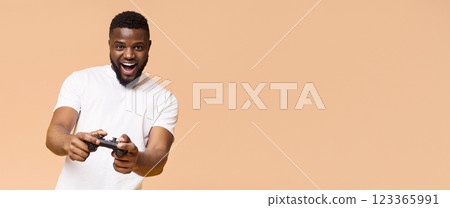 A young African American man wearing a white T-shirt is smiling excitedly as he plays video games. He is holding a black controller in both hands, pointing it towards the camera, copy space A young African American man wearing a white T-shirt is smiling excitedly as he plays video games. He is holding a black controller in both hands, pointing it towards the camera, copy space 123365991