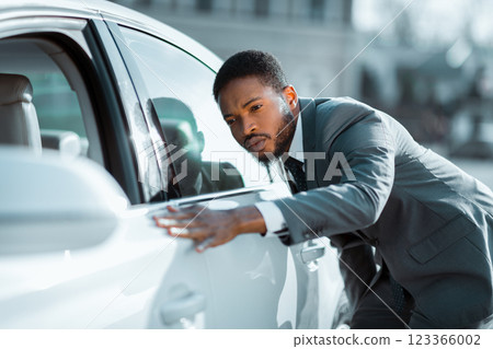 African American man in a suit is inspecting a white car during the day. He has his hand on the car door and is looking at the paint job. The car is parked on a street. 123366002
