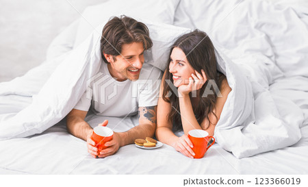 A couple, a man and a woman, are lying in bed, covered by white bedding. They are both smiling and looking at each other, holding mugs of coffee and a plate of cookies. A couple, a man and a woman, are lying in bed, covered by white bedding. They are both smiling and looking at each other, holding mugs of coffee and a plate of cookies. 123366019