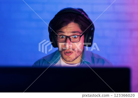 A young man with a shocked expression is seen staring intently at his computer screen while wearing headphones. The room is dimly lit with blue and purple hues, suggesting a late-night gaming session. A young man with a shocked expression is seen staring intently at his computer screen while wearing headphones. The room is dimly lit with blue and purple hues, suggesting a late-night gaming session. 123366020