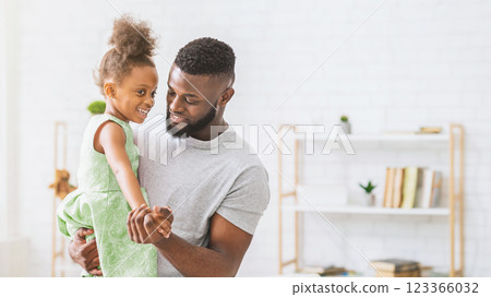 African American father smiles and holds his daughter in his arms, both looking at each other. The background features a bookshelf with books and decor in a white home interior. 123366032