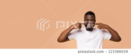African American man wearing a white t-shirt is standing against a beige backdrop. He is smiling widely and pointing to his teeth with both index fingers, copy space African American man wearing a white t-shirt is standing against a beige backdrop. He is smiling widely and pointing to his teeth with both index fingers, copy space 123366039