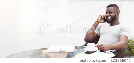 A man sits at a table, smiling and talking on his phone while holding a cup of coffee. 123366043