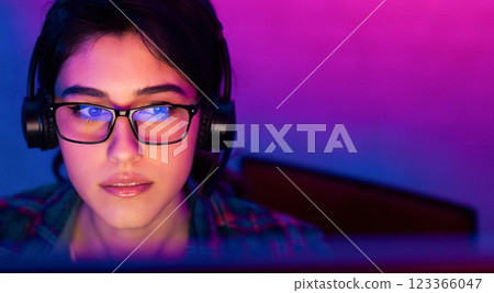 A close-up shot of a young woman with dark hair wearing glasses and headphones, looking intently at a computer screen. The background is a vibrant mix of purple and blue lighting, copy space A close-up shot of a young woman with dark hair wearing glasses and headphones, looking intently at a computer screen. The background is a vibrant mix of purple and blue lighting, copy space 123366047