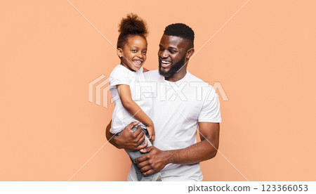 A smiling African American father holds his happy daughter in his arms while they both look at the camera. The photo has a peach colored background. A smiling African American father holds his happy daughter in his arms while they both look at the camera. The photo has a peach colored background. 123366053