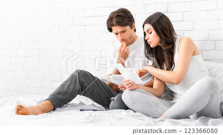 A young couple sits together on a white bed, looking at bills and documents. The man, wearing a white shirt and grey pants, is biting his fingernail and appears stressed. 123366080