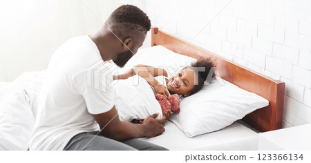 Black father sits on the edge of a bed, tucking in his little daughter who is smiling and holding a stuffed animal. The room is bright and the bed is neatly made with white sheets and pillows. 123366134