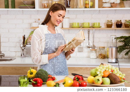 A woman stands in her kitchen wearing an apron and reading a cookbook. She is preparing to cook a meal, as evidenced by the fresh produce on the counter. 123366180