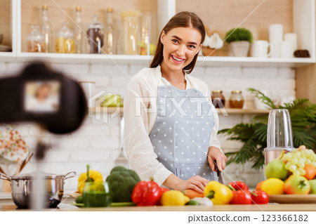 A woman in a kitchen, wearing an apron and smiling at the camera, prepares a meal while filming for a cooking show. 123366182