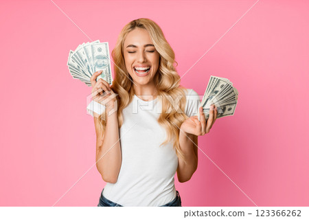A young woman with blonde hair and a white t-shirt is smiling excitedly while holding US dollar bills in both hands. She is standing against a pink background, looking happy and successful. 123366262