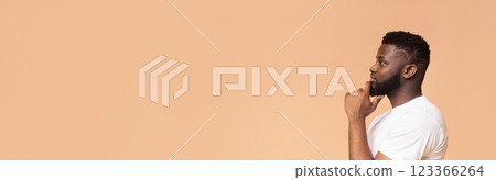 This image features a young African American man thinking while looking to the side. He is wearing a white t-shirt and standing against a peach-colored background, copy space 123366264