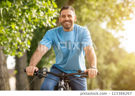 A man smiles while riding a bicycle on a sunny day in a park. The background is filled with green trees and foliage. The man is wearing a blue t-shirt and jeans 123366267