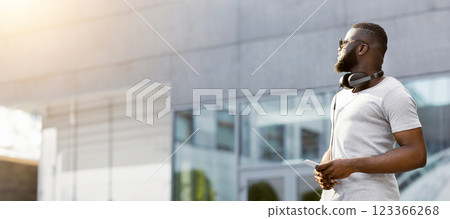 A man with headphones around his neck looks up at a city building, with a phone in his hand. A man with headphones around his neck looks up at a city building, with a phone in his hand. 123366268