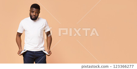A young black man with a beard is standing against a beige backdrop. He is wearing a white t-shirt and blue jeans and is checking his pockets, which appear to be empty, copy space 123366277