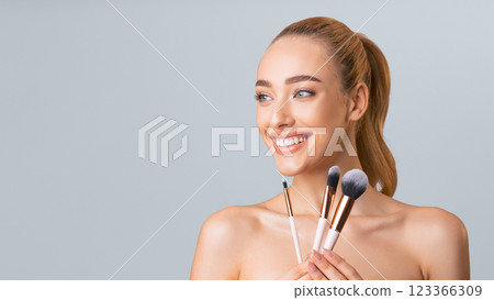 A young woman with long blonde hair pulled back into a ponytail is holding three makeup brushes in front of a light grey background. She is smiling and looking at copy space 123366309