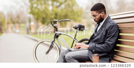 A business professional black man uses his laptop on a park bench beside his bike. The scene is calm and modern, highlighting a balance of work and relaxation in nature. 123366320