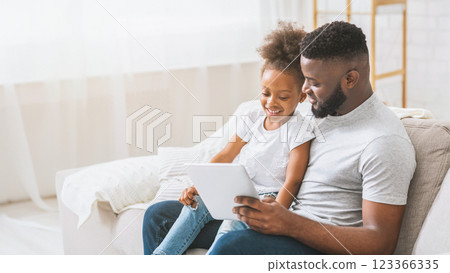 Black father and daughter sit on a couch together in their home, looking at a tablet. The daughter is sitting on the fathers lap, and they both appear to be enjoying the content on the tablet 123366335