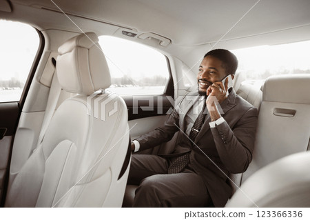 Black businessman in a suit sits in the backseat of a car, talking on his phone. He is smiling and looking out the window. The car appears to be a luxury vehicle with leather seats. 123366336