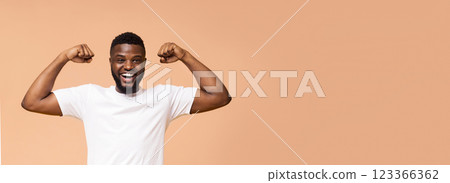 A happy, young, Black man with a short beard is flexing his muscles while standing in front of a beige background. He is wearing a white t-shirt and has a big smile on his face, copy space, panorama 123366362