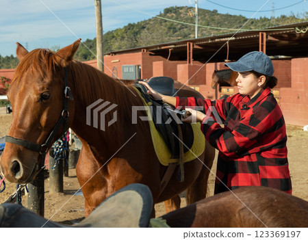 Positive young woman invites to ride horse at horse farm Positive young woman invites to ride horse at horse farm 123369197