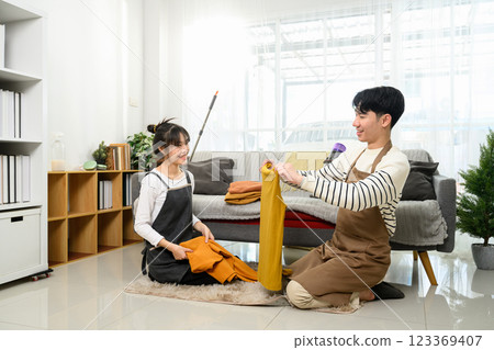 Happy young couple wearing aprons folding laundry and tidying up their living room Happy young couple wearing aprons folding laundry and tidying up their living room 123369407
