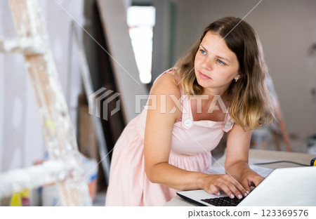Young attractive girl checks the quality and correctness of the completed repairs using a laptop inside cottage Young attractive girl checks the quality and correctness of the completed repairs using a laptop inside cottage 123369576