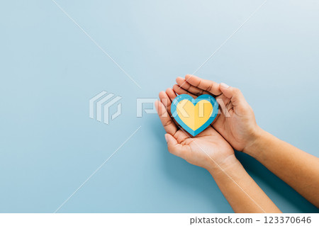 A yellow and blue heart held by hands on a yellow background represents Down Syndrome 123370646