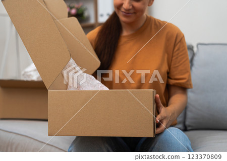 E-Commerce Elation: A delighted woman, on her couch, unboxes a carton parcel from an online order, celebrating the joy of shopping online.  123370809