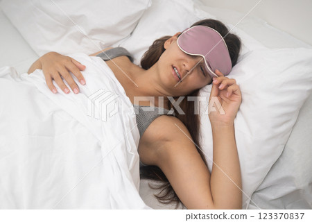 A vexed woman in a sleep mask awakens, despising the alarm's intrusion, sleep-deprived, captured in a close-up moment.  123370837