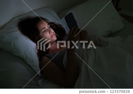 A woman, in the dark of night, lies in bed, fixated on her phone's glow, image portraying the struggles of insomnia and smartphone addiction. A woman, in the dark of night, lies in bed, fixated on her phone's glow, image portraying the struggles of insomnia and smartphone addiction. 123370839