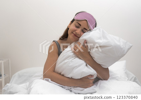 In the bedroom's morning glow, an enchanting woman with a sleep mask grins, holding a plush white pillow. In the bedroom's morning glow, an enchanting woman with a sleep mask grins, holding a plush white pillow. 123370840
