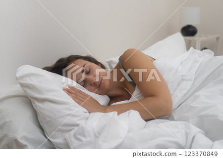 Serenely asleep, a young lady rests under a white blanket on her comfortable orthopedic mattress in a well-lit bedroom. 123370849
