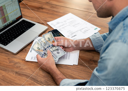 Man counting dollar bills while reviewing utility bills and household expenses. Budget planning and financial management at home. Personal finance and tax calculation concept. Man counting dollar bills while reviewing utility bills and household expenses. Budget planning and financial management at home. Personal finance and tax calculation concept. 123370875