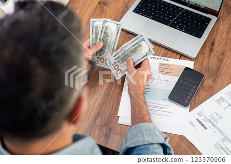 Grey haired man counts one hundred dollars banknotes over utility bills sitting at table with laptop, mature businessperson prepares to pay monthly household expenses with cash Grey haired man counts one hundred dollars banknotes over utility bills sitting at table with laptop, mature businessperson prepares to pay monthly household expenses with cash 123370906