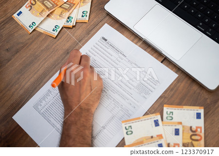 Corporate executive sits at table with euro bills and laptop with pen in hand completing 1040 form to report salary bonuses and deductions determining tax Corporate executive sits at table with euro bills and laptop with pen in hand completing 1040 form to report salary bonuses and deductions determining tax 123370917