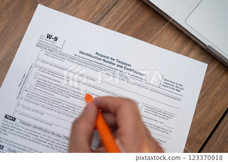 Real estate agent meticulously completes W-9 form ensuring TIN correctly provided for reporting commissions from property sales at kitchen table holding pen with laptop nearby 123370918