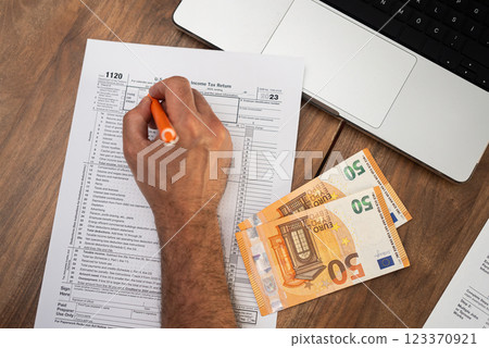 Tax advisor meticulously fills out 1120 tax form on table using laptop and referring to euro bills to ensure accurate currency conversions and reporting 123370921