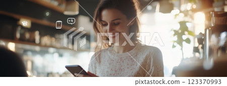 Cheerful young woman happily using her smartphone in a sunny cafe. With social media icons floating above her head Cheerful young woman happily using her smartphone in a sunny cafe. With social media icons floating above her head 123370993