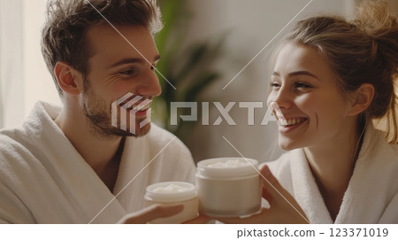 Smiling man and woman wearing bathrobes are holding opened jars of cosmetic cream, enjoying their skincare routine together in a cozy home atmosphere 123371019