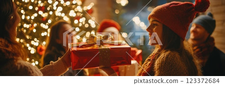 Woman exchanging a beautifully wrapped Christmas gift with a friend by the tree. A festive moment of giving during the holidays. 123372684