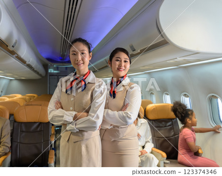 Two smiling flight attendants standing in commercial aircraft cabin while passengers sitting by window Two smiling flight attendants standing in commercial aircraft cabin while passengers sitting by window 123374346