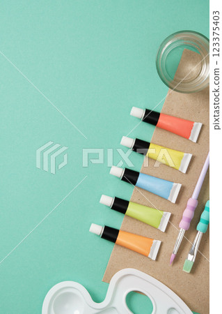 Preparing to painting. Tubes of acrylic paint, brushes, a glass of water on mint green background. Place for your text. 123375403