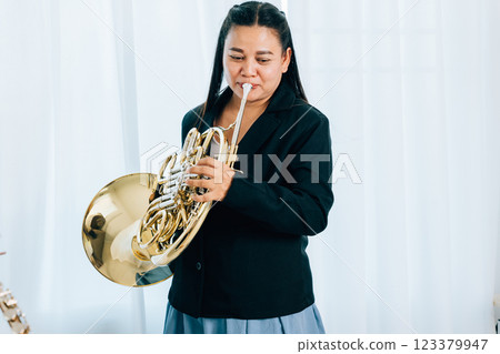 Standing gracefully in a cozy living room, a female musician plays a shiny gold horn. The soft curtain background enhances the warm and artistic atmosphere of the scene. 123379947