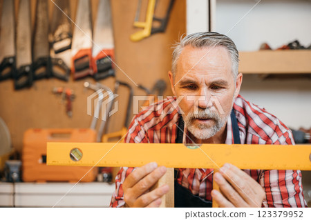 A senior carpenter in a red plaid shirt carefully uses level to measure wooden stool, working precisely in well-equipped woodworking shop filled with various tools, showcasing his skill in carpentry 123379952
