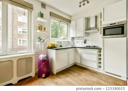 This contemporary kitchen features sleek white cabinetry, minimalistic decor, and large windows allowing natural light to fill the space. A vibrant purple fridge adds a pop of color. 123380015